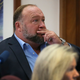 Alex Jones sits with his two fingers to his mouth, staring off to the right.